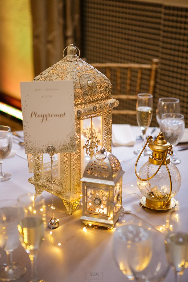 a table topped with lots of glasses and plates covered in gold colored decorations next to a lit lantern