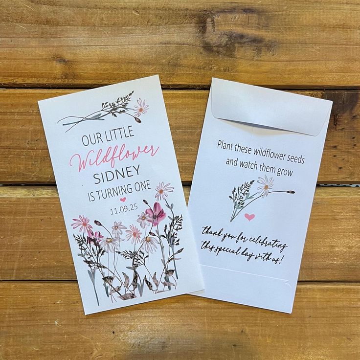 two wedding cards with flowers on them sitting next to each other in front of a wooden background