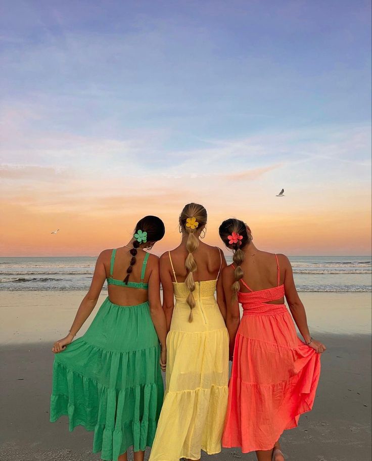 three women standing on the beach looking out at the ocean with their backs to the camera