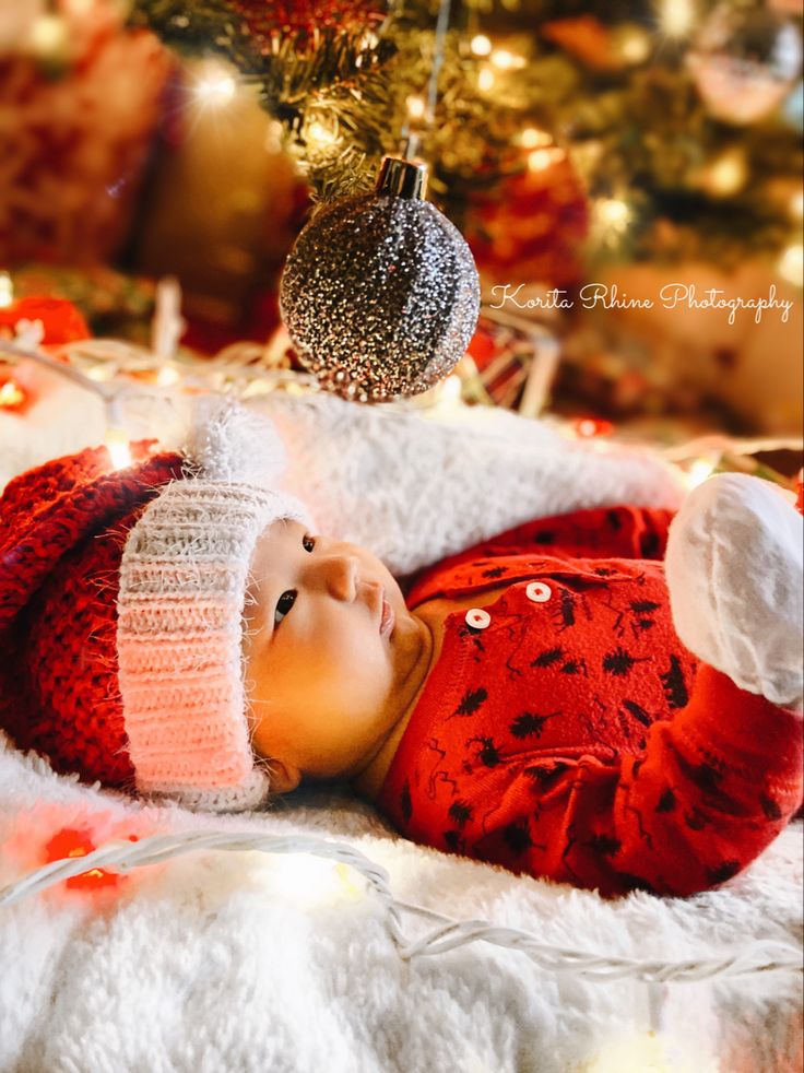 a baby wearing a red and white hat laying in front of a christmas tree