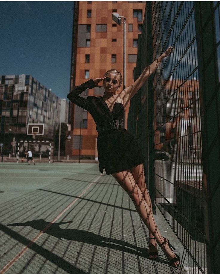 a woman leaning against a fence with her arms outstretched and legs crossed, in front of a tall building