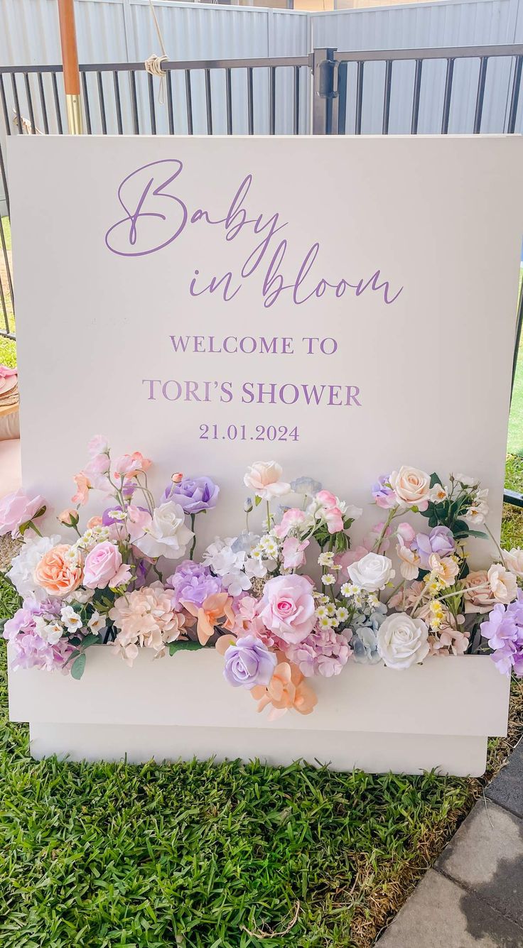 a welcome sign with flowers on it in front of a fence and grass area at a baby shower