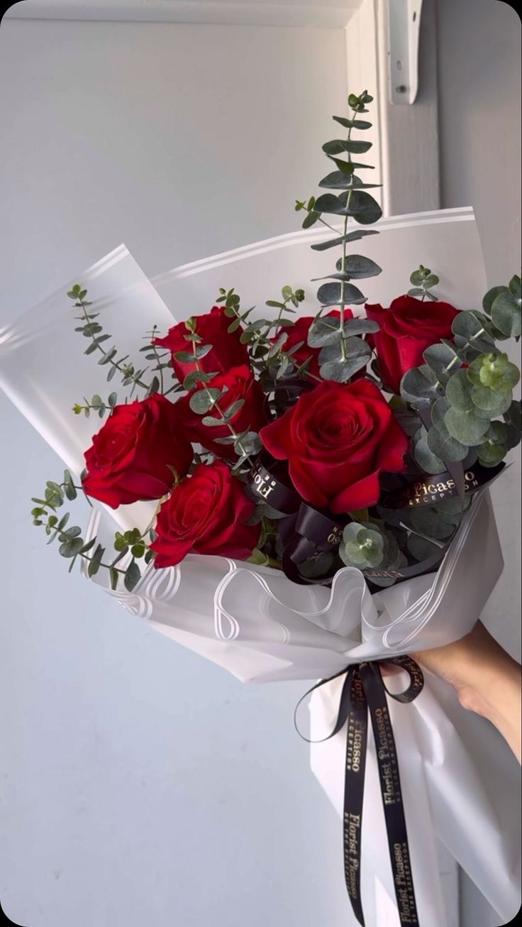 a bouquet of red roses wrapped in white paper with greenery and ribbon tied around it