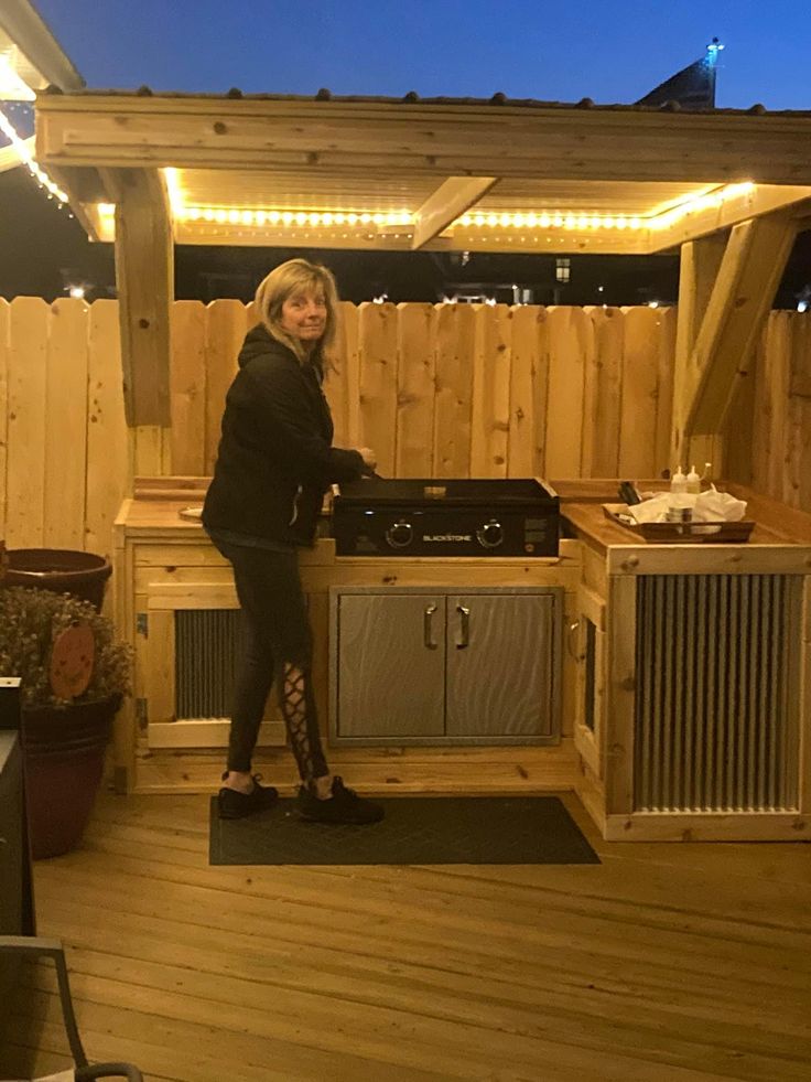 a woman standing in front of an outdoor kitchen with lights on the roof and wood flooring