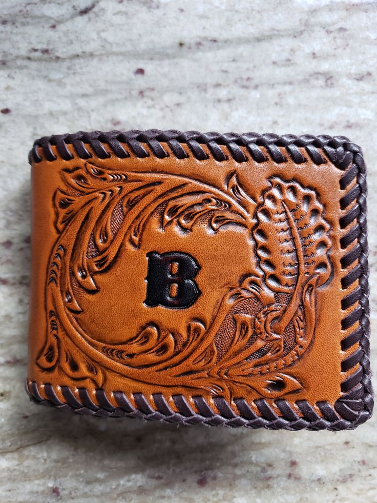 This wallet is tooled using genuine vegetable tanned cowhide and hand laced with calfskin lace. The carving and tooling is done in the Sheridan Style with a rich color stain applied overall. A top finish has been applied to protect your wallet for years of wear. The inside has six pockets for credit cards and I D. A name or initials may be added at no extra charge. Wallet can also be hand saddle stitched upon request. Hand Tooled Leather Wallet, Traditional Brown Leather Wallet, Custom Hand Tooled Leather Trifold Wallet, Hand Tooled Bifold Wallet, Artisan Hand Tooled Leather Wallet, Artisan Hand Tooled Leather Trifold Wallet, Custom Hand-tooled Trifold Wallet, Artisan Hand Tooled Bifold Wallet, Rectangular Leather Wallet With Hand Tooling