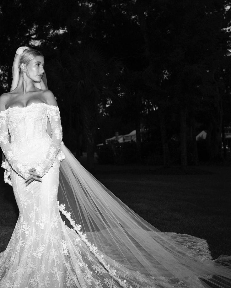 a woman wearing a wedding dress and veil standing in the grass with trees behind her