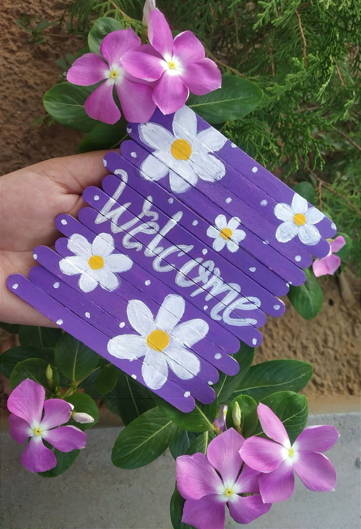 purple and white flowers are being held up by someone's hand with the word welcome painted on it