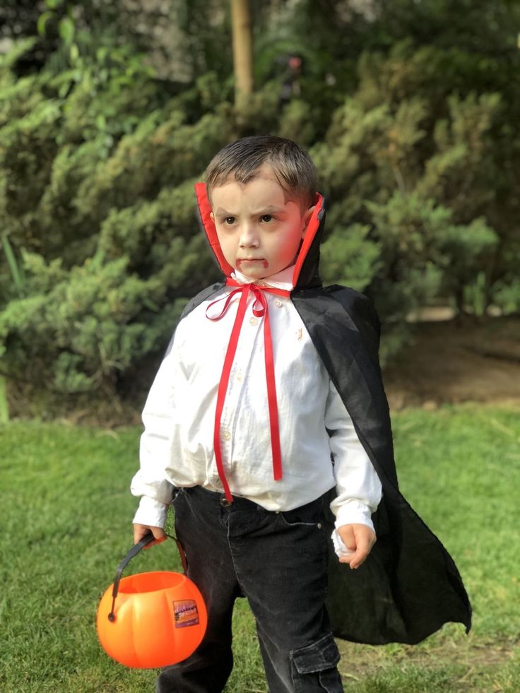 a young boy dressed up as a vampire holding an orange bucket and wearing a black cape