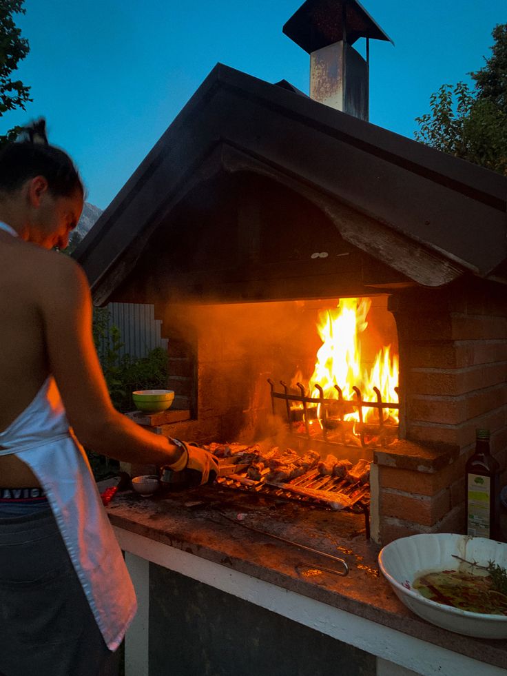 a woman is cooking food on an outdoor grill with flames in the background at night