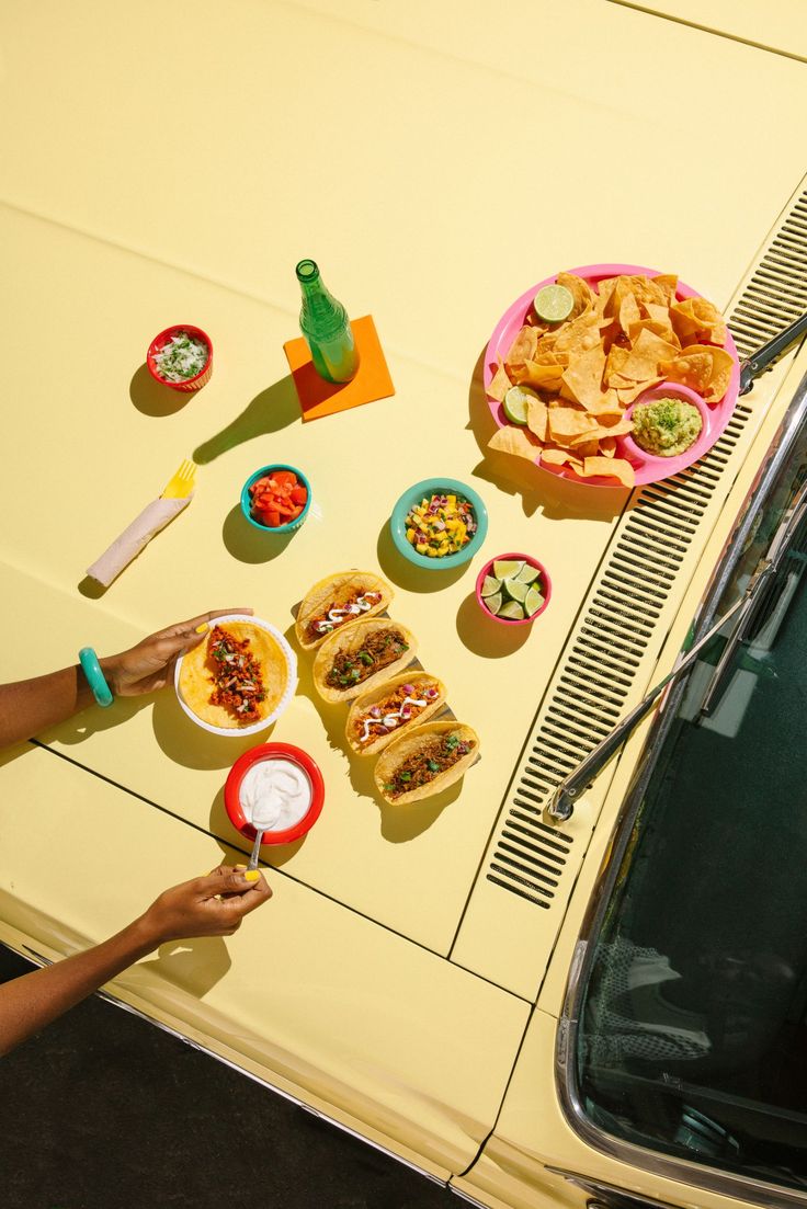 people are eating food out of plates on the hood of a yellow car, while another person stands next to them