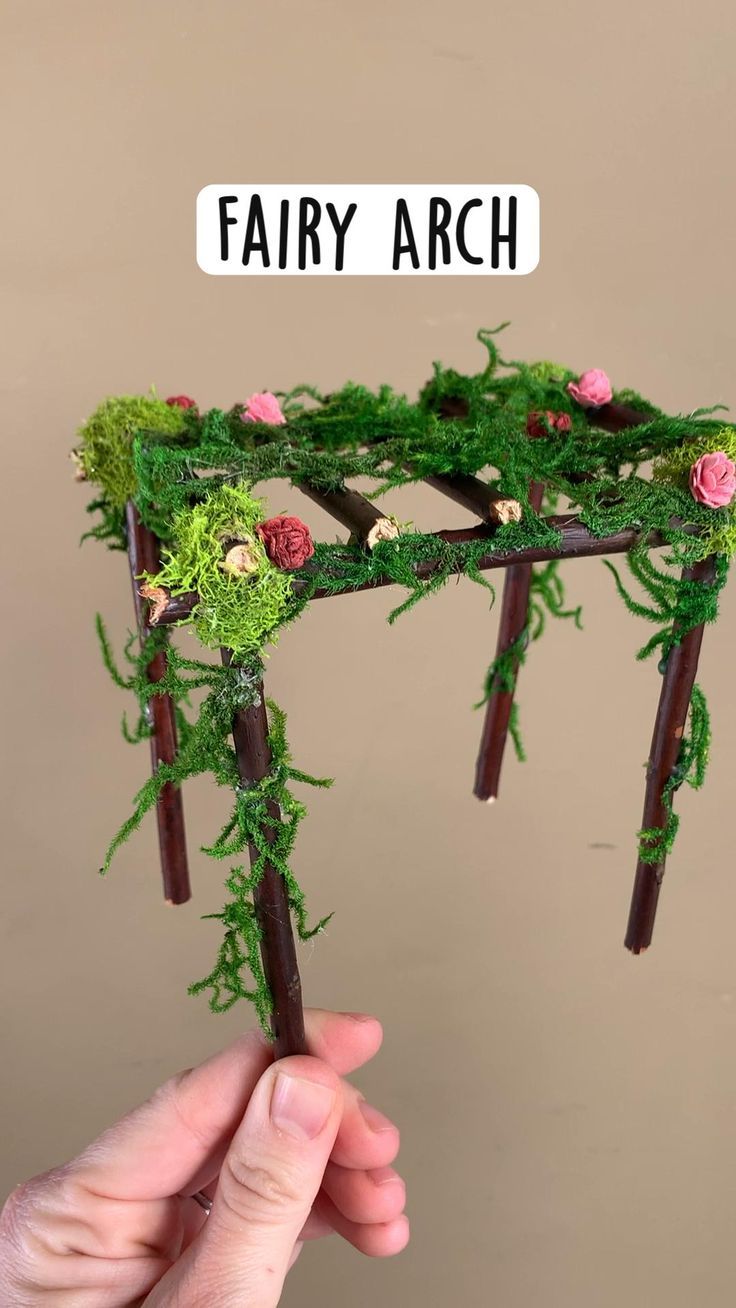 a hand holding up a fake fairy arch made out of branches and moss with flowers on it