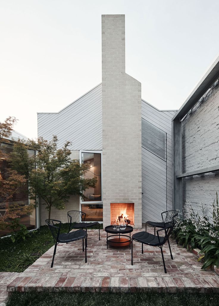 an outdoor fireplace in the middle of a brick patio with chairs around it and plants on either side