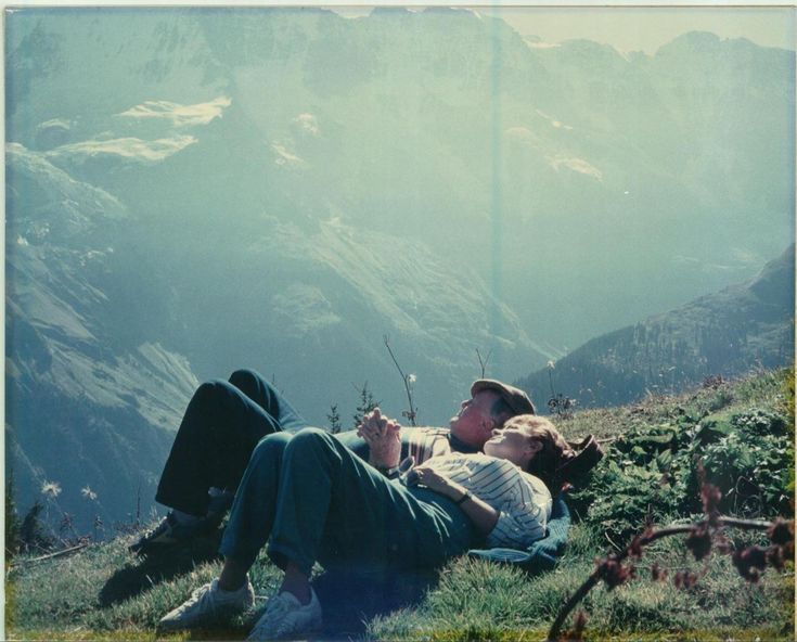 two people are laying on the grass in front of some mountains and looking at each other