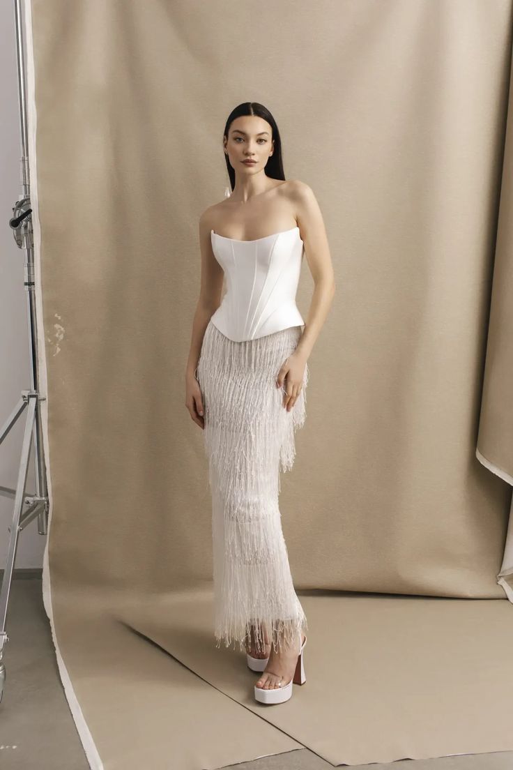 a woman standing in front of a backdrop wearing a white top and fringed skirt