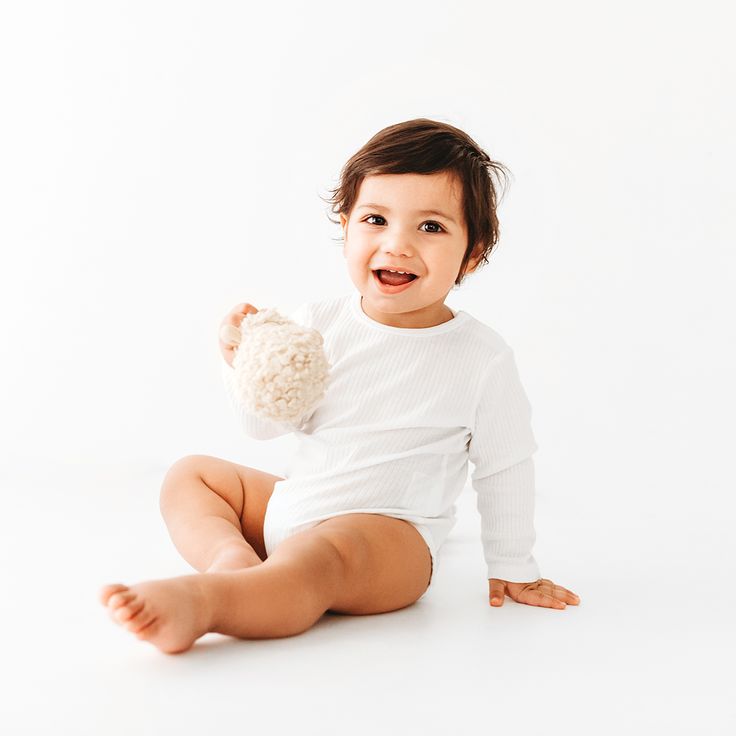 a smiling baby sitting on the floor with a stuffed animal in his hand and wearing a white bodysuit