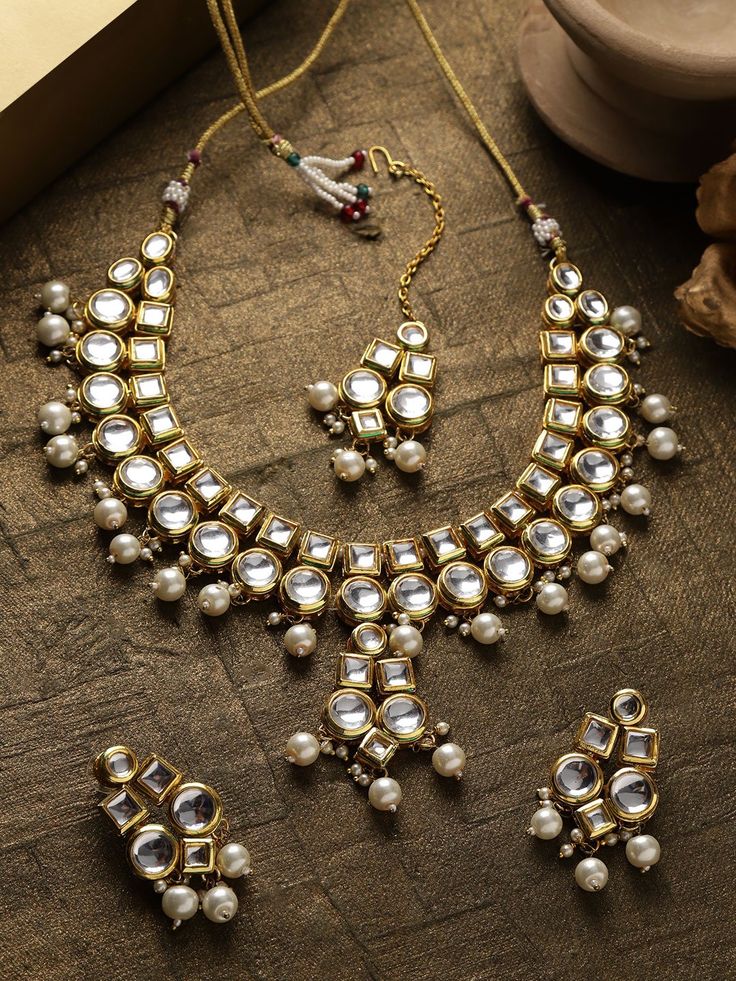 This stunning jewelry set consists of a classic drop necklace, a maang tikka, and a pair of matching drop earrings. The gold toned & off-white pearl necklace with a central drop shown here comes with kundan stone studs & beads, is gold-plated, and secured with an adjustable drawstring closure. A pair of matching drop earrings come secured with a post and back closure. This beautiful statement piece can add impact to any outfit for any festive occasion! Product color may vary based on the monitor Bollywood Kundan Bridal Necklace With Pearl Drop, Bollywood Style White Kundan Necklace With Pearl Drop, Silver Kundan Necklace With Pearl Drop, Kundan Necklace With Pearl Drop For Celebrations, Festive White Kundan Necklace With Pearl Drop, White Kundan Necklace With Mirror Work, White Kundan Jeweled Jewelry, White Kundan Jewelry With Pearl Drop, Kundan Jewelry Sets With Pearl Drop As Gift