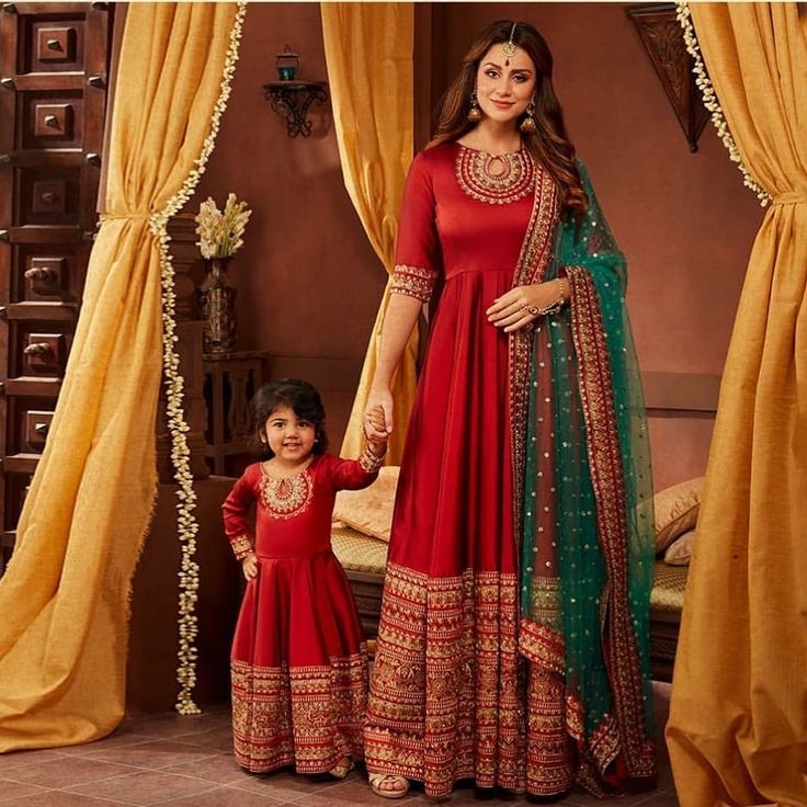 a woman and child standing in front of a window wearing red dresses with green dupatta