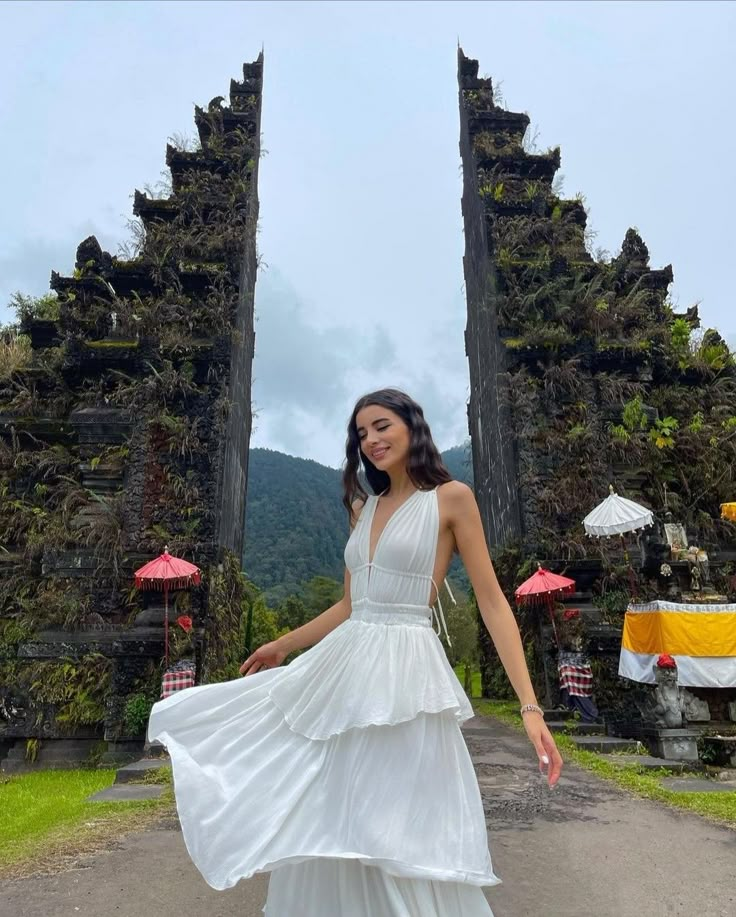 a woman standing in front of two stone gates wearing a white dress and holding an umbrella