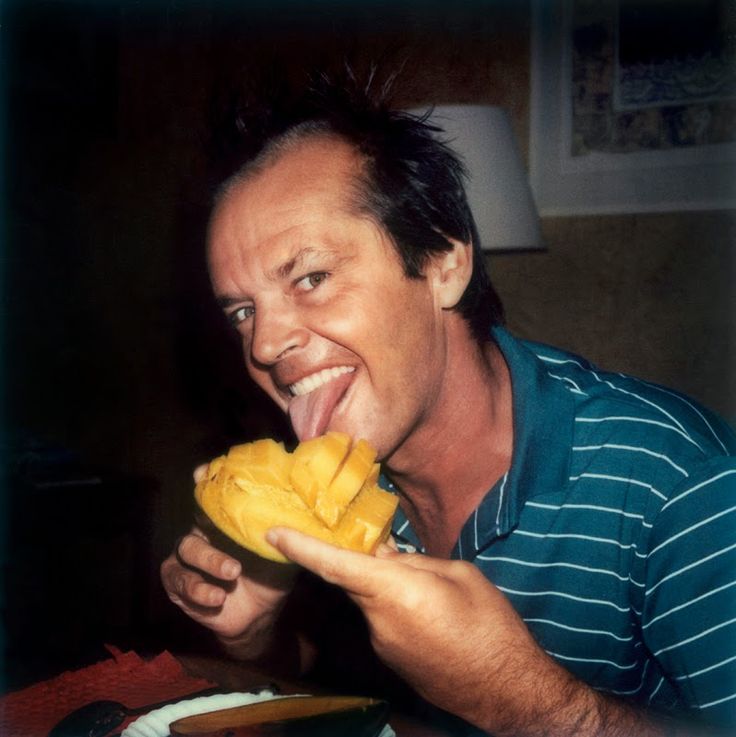 a man is smiling and eating a mango fruit while sitting at a table in front of him