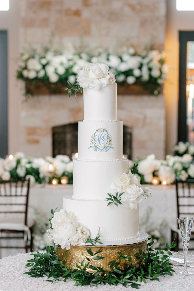 a wedding cake with white flowers and greenery on the table in front of a fireplace