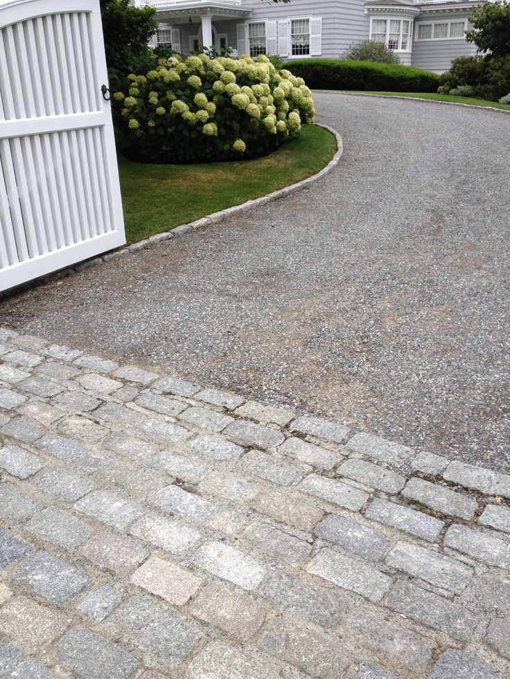 the driveway is made of cobblestones and has a white gate on one side