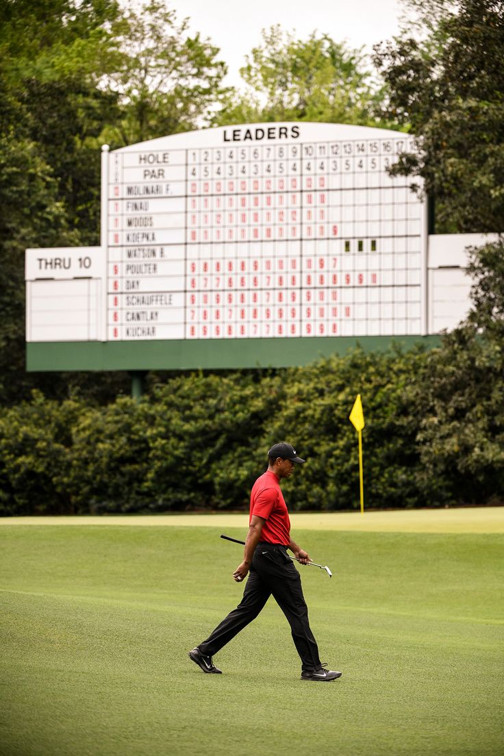 a man walking across a green covered golf course with a scoreboard in the background