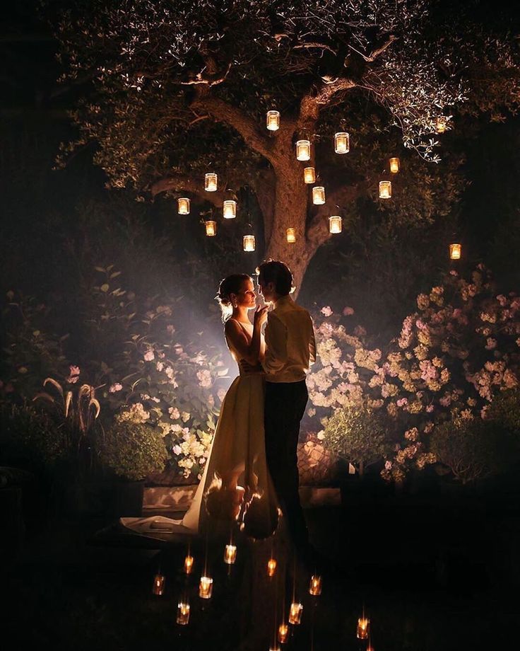 a man and woman standing under a tree with candles in the dark, surrounded by flowers