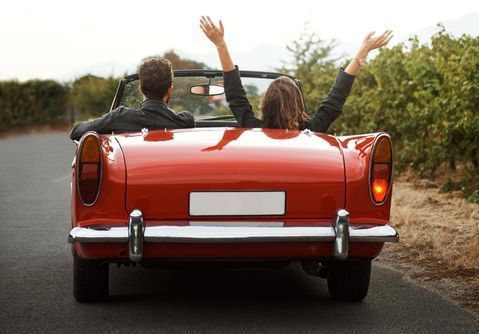 two people sitting in the back of a red convertible car with their hands up to the sky