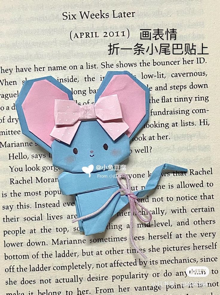 an origami mouse with pink ears and bow on it's head sitting in front of a book