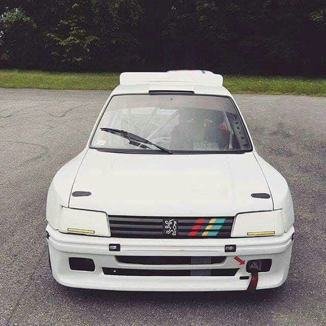 the front end of a white car parked in a parking lot with trees in the background