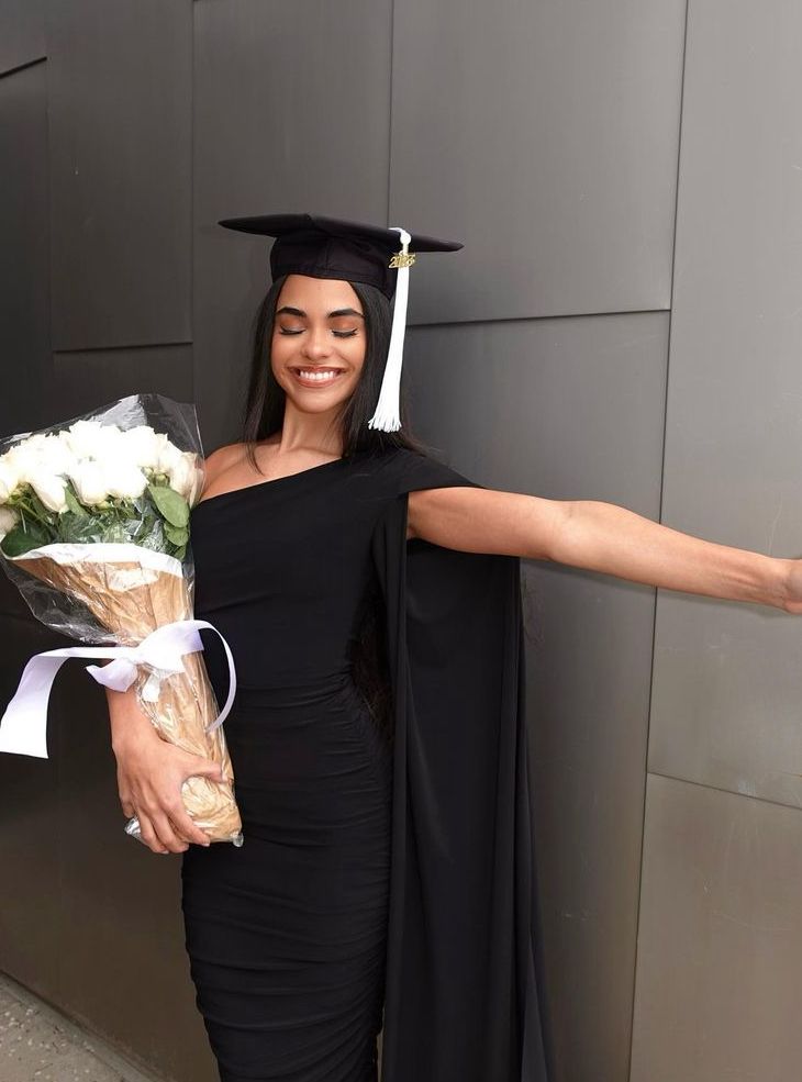 a woman in a graduation cap and gown holds flowers while standing next to a wall