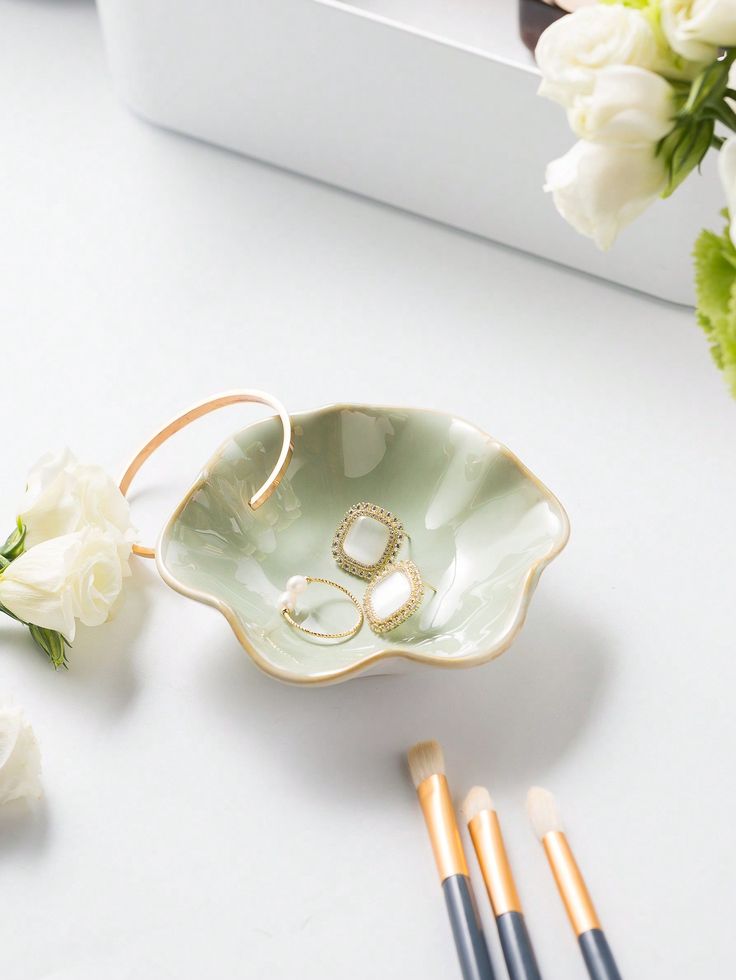 wedding rings in a flower shaped bowl next to makeup brushes and flowers on a table