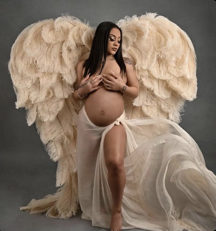 a pregnant woman with angel wings posing for a photo in front of a gray background