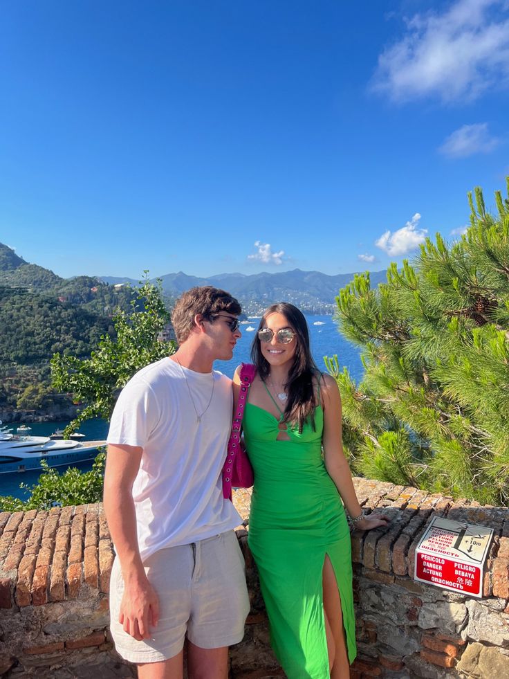 a man and woman standing next to each other on top of a stone wall near the ocean