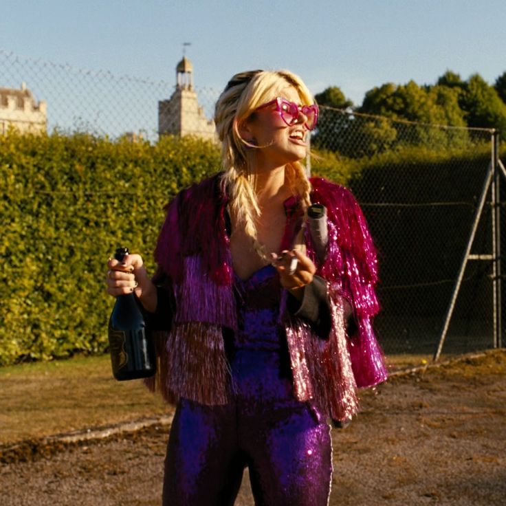 a woman in purple and pink outfit holding a wine bottle while standing next to a fence