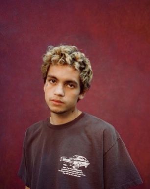 a man with curly hair standing in front of a red wall wearing a black shirt