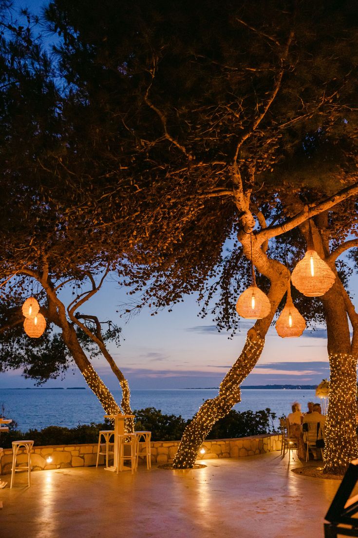 an outdoor dining area with lanterns hanging from the trees and lights on the tables below