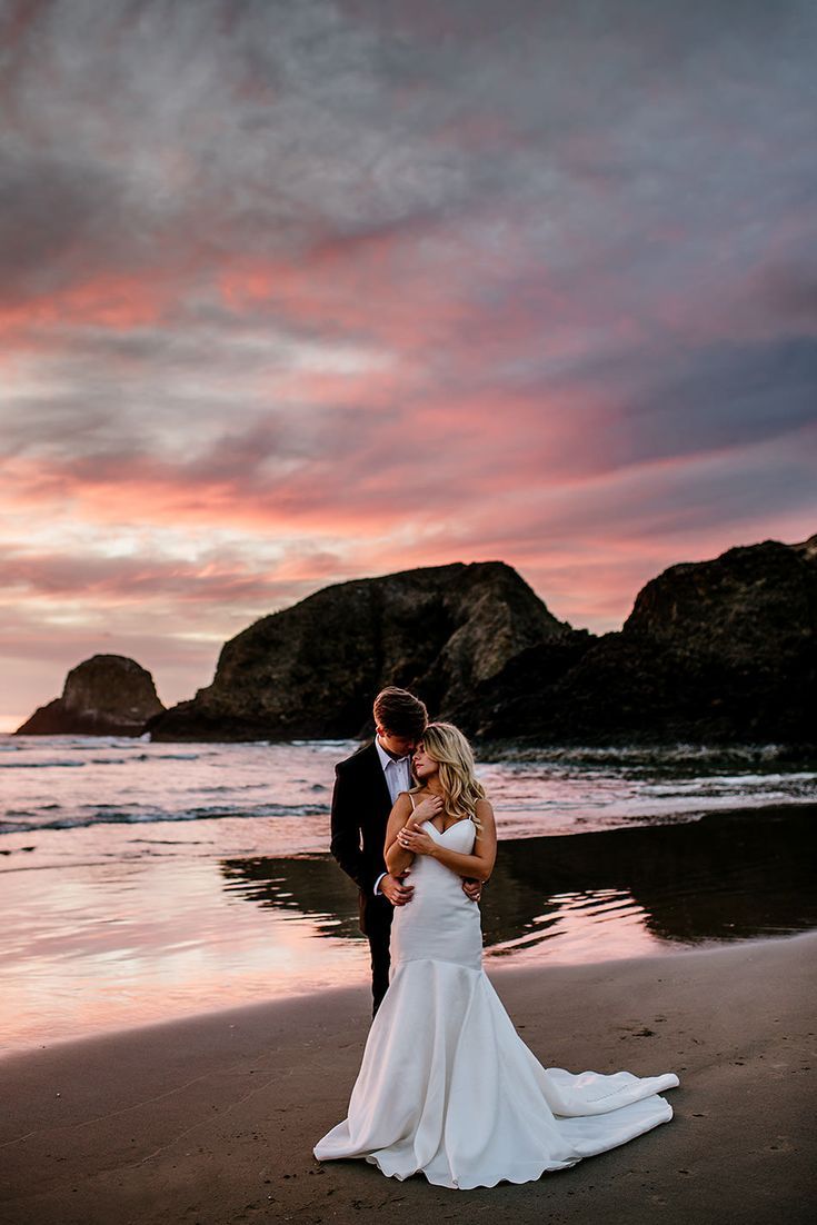 a bride and groom standing on the beach in front of an ocean sunset with pink clouds