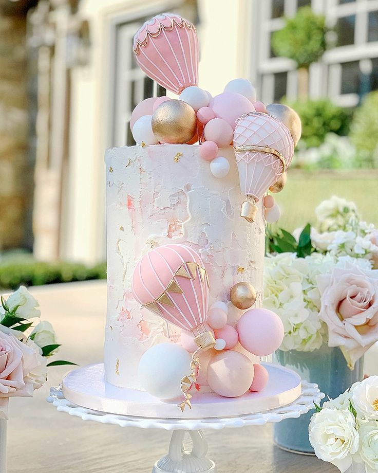 a white and pink cake with balloons on it sitting on a table next to flowers