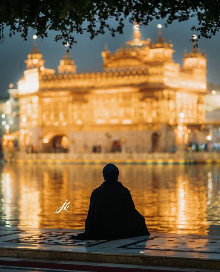 a man sitting on the ground in front of a large building with lights reflecting off it's water