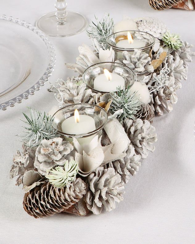 pine cones and candles are arranged on a white table cloth with silver rimmed glasses