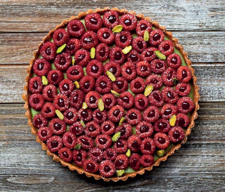 a tart with raspberries and pistachios on top, sitting on a wooden table