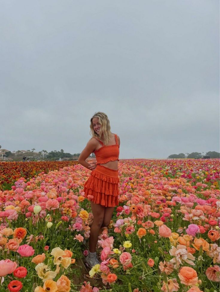 a woman in an orange dress standing in a field full of flowers with her hands on her hips