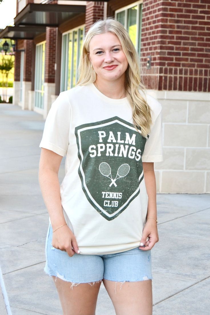 a woman standing on the sidewalk with her tennis racket in her hand and wearing a shirt that says palm springs tennis club