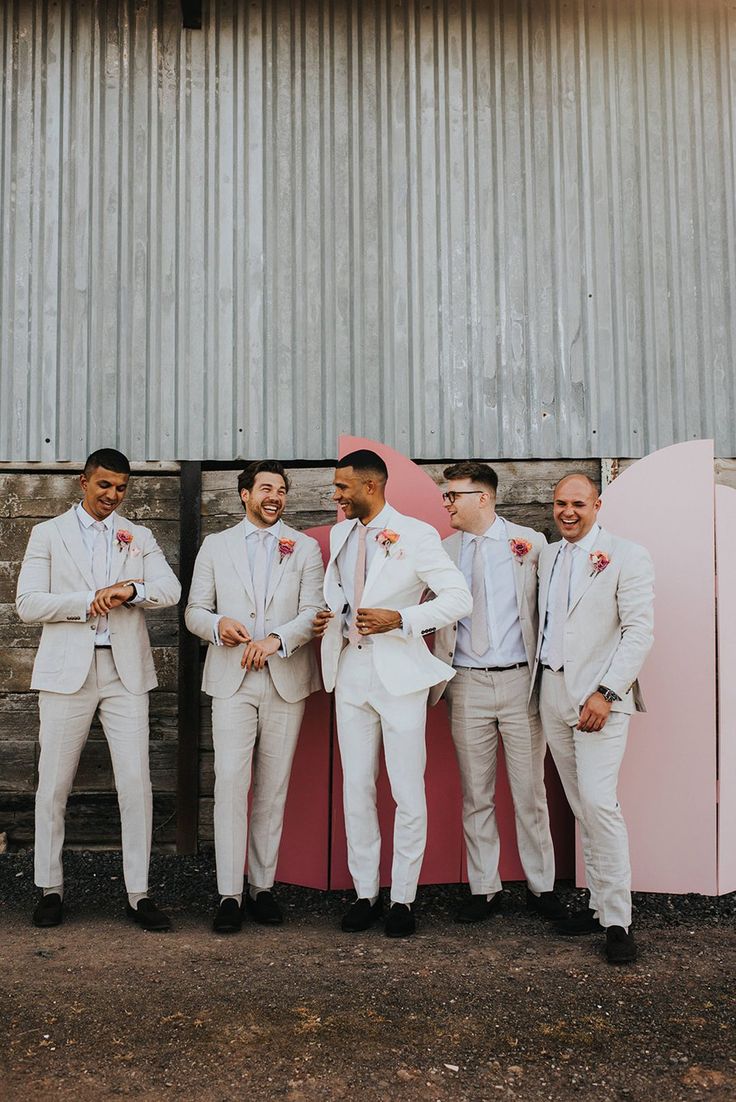 Groom in a white suit and groomsmen in cream suits with pink ties and pink flower buttonholes in front of the pink wedding screen background Light Grooms Suit, Bright Wedding Groomsmen, White And Pink Mens Suit, Groom Blush Wedding, Blush Pink Groomsmen Suit, White Suit With Pink Tie, Pink Grooms Suit, Coloured Wedding Suit, Pink Wedding Bride And Groom