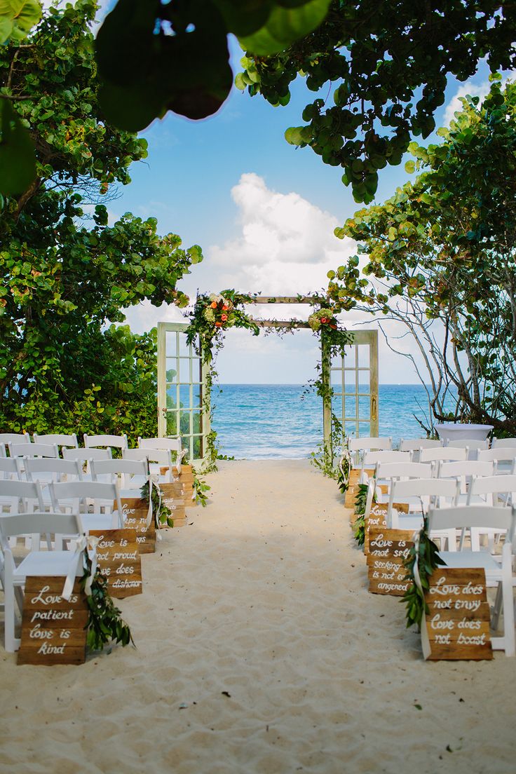 an outdoor ceremony set up on the beach with white chairs and greenery at the end