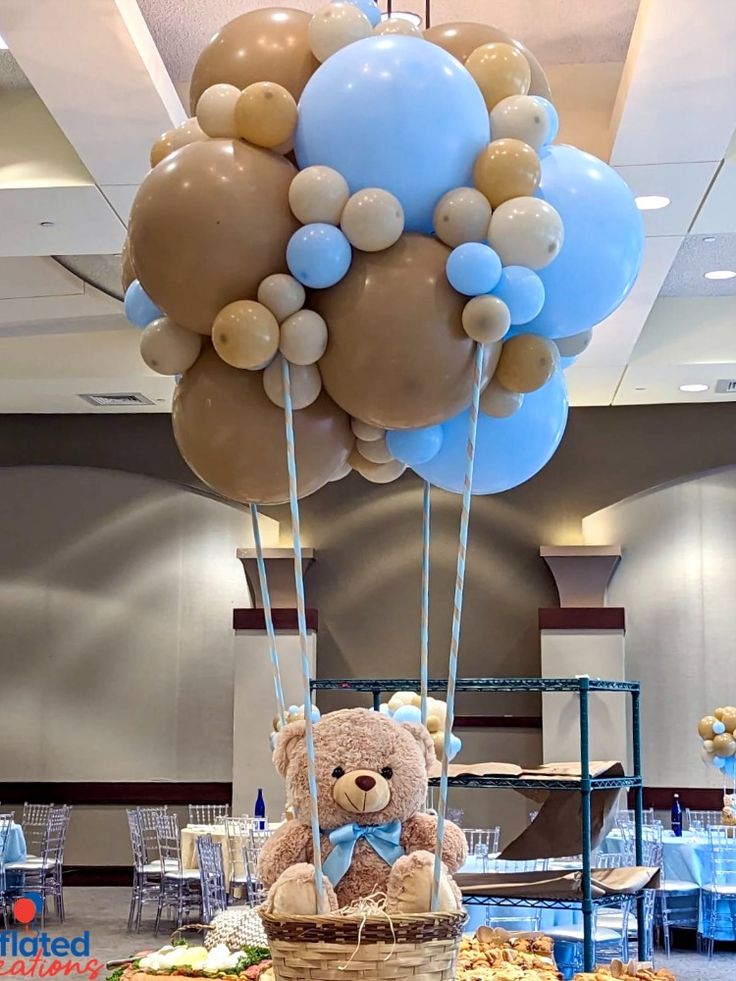 a teddy bear sitting on top of a basket filled with blue and gold balloons in front of a table