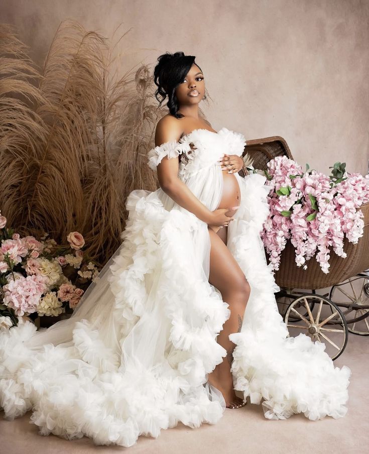 a pregnant woman in a white gown posing for a photo with flowers and a wagon behind her