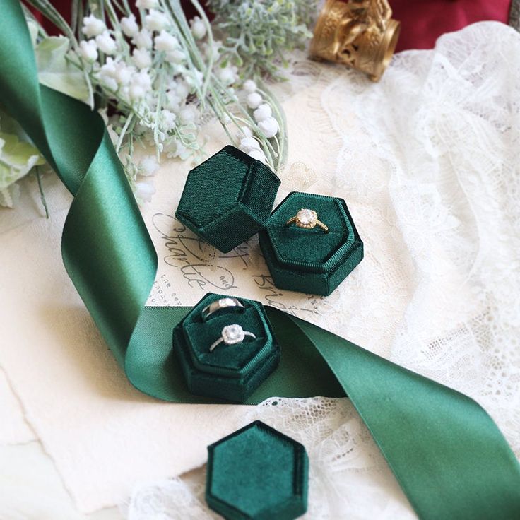 three green velvet ring boxes sitting on top of a lace table cloth next to flowers