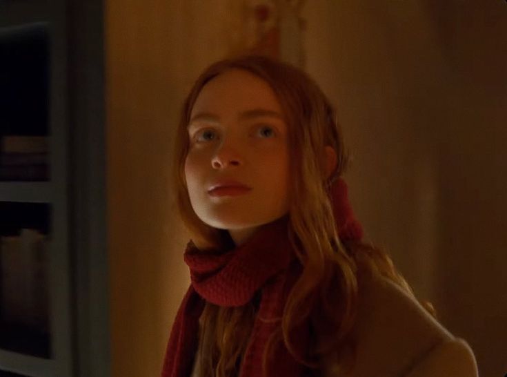 a woman with long red hair wearing a scarf and looking at the camera in a dimly lit room